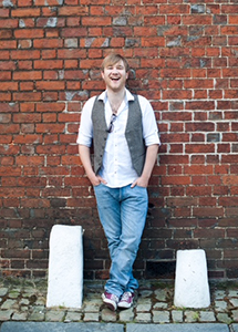 A smiling man in a white shirt, grey vest, and blue jeans leans against a red brick wall.