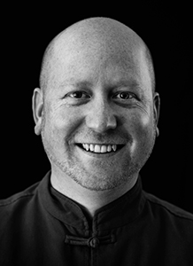 Black and white headshot of a smiling, bald man wearing a dark mandarin collar shirt against a black background.