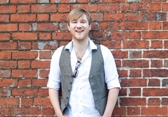 A smiling man with light hair wearing a white shirt and grey vest against a red brick wall.