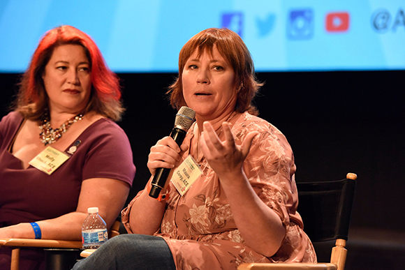 A woman in a pink floral shirt speaks into a microphone while another woman watches.