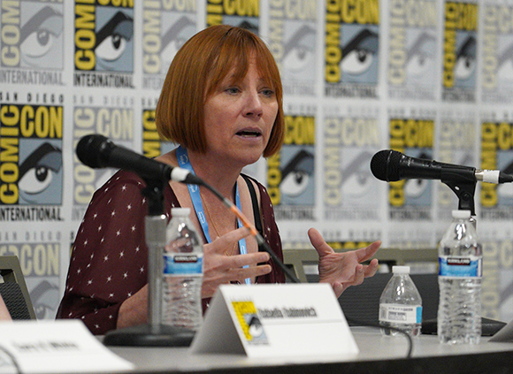 A woman with reddish-brown hair speaks into a microphone at a Comic-Con panel.