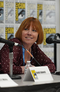 A woman with shoulder-length reddish hair smiles behind a microphone at a Comic-Con event.