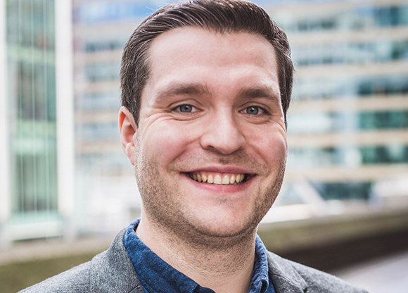 A smiling man with short brown hair wearing a grey blazer and blue shirt against a blurred building background.
