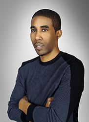 A man with short dark hair and a dark long-sleeved shirt posing with arms crossed against a grey background.