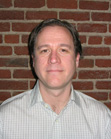 A man with short brown hair wearing a light checkered shirt, standing in front of a red brick wall.