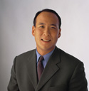 A smiling man wearing a dark suit, blue shirt, and dark tie against a light background.