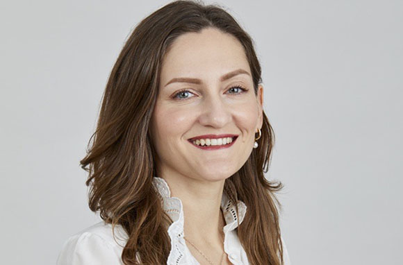 A smiling woman with wavy brown hair wearing a white ruffled blouse against a light gray background.