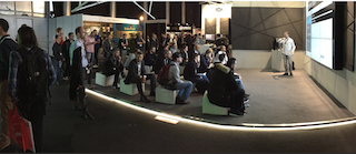 A man in a white shirt presents to a seated audience in a large exhibition hall.