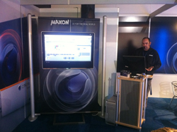 A man stands next to a large screen displaying Maxon software, with a computer and monitor on a stand.