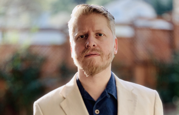 A man with blonde hair and a beard wearing a white blazer and a dark blue shirt.