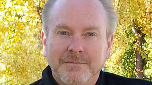 A man with light hair and a goatee looks forward against a backdrop of bright yellow autumn foliage.