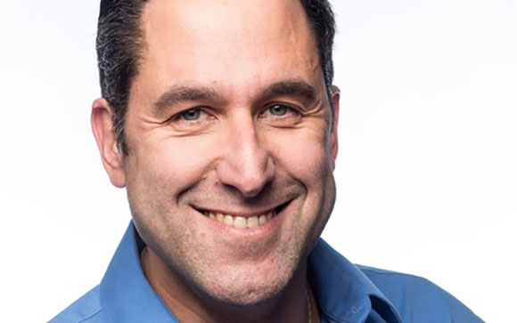 A smiling man with short dark hair wearing a blue button-down shirt against a white background.