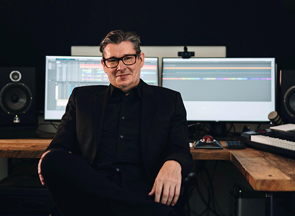 A man wearing glasses and a black suit sits in front of computer monitors in a music studio.