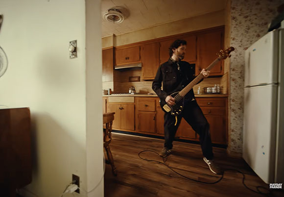 A man in black clothing plays an electric bass guitar in a kitchen with wooden cabinets.