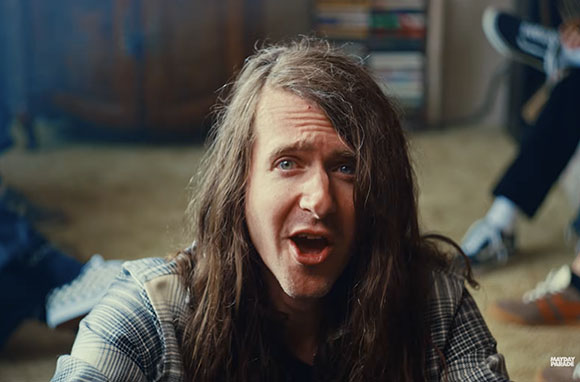 A man with long brown hair and an expressive face looks directly at the camera.