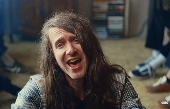 A man with long, messy brown hair smiles widely with his mouth open.