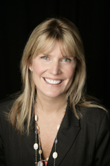 A smiling woman with blonde hair and bangs wearing a dark blazer and a beaded necklace against a black background.