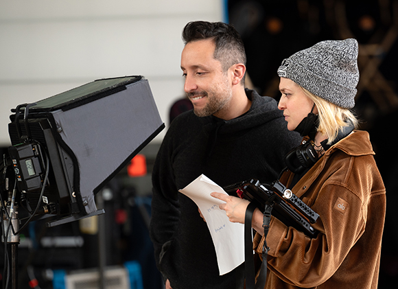 A man in a black hoodie and a woman in a grey beanie look toward a large studio light on a film set.