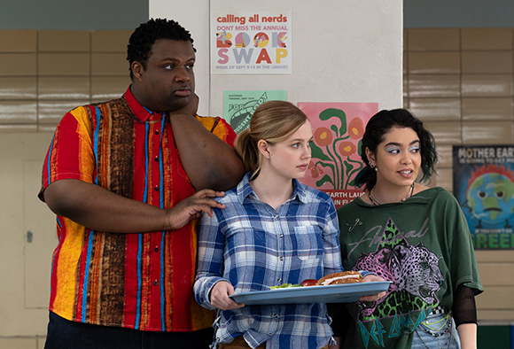 A man in a colorful shirt and two young women stand in a hallway near a pink poster.