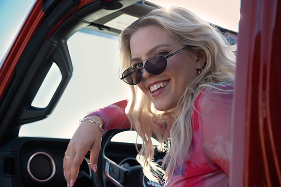 A smiling blonde woman in sunglasses leans against the open door of a red vehicle.