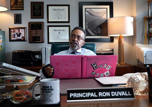 A man with glasses and a beard sits at a desk behind a pink book and a nameplate reading Principal Ron Duvall.