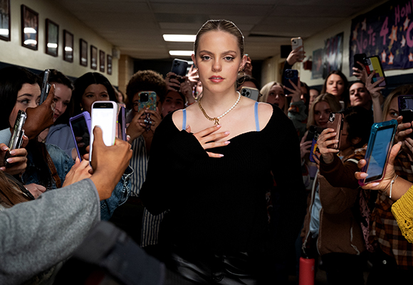 A young woman in a black top stands in a crowded hallway as people hold up phones to take her picture.
