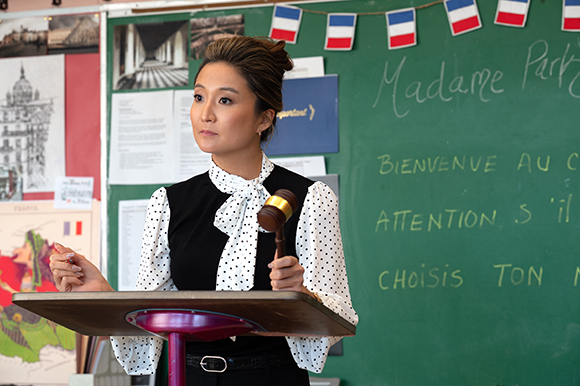 A woman in a polka-dot blouse holds a wooden gavel while standing behind a podium in a classroom.