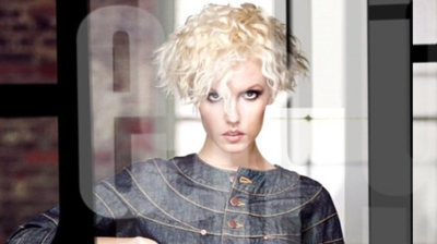 A woman with short, curly blonde hair wearing a denim top, framed by large translucent letters.
