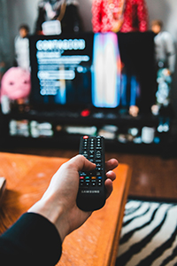 A hand holds a black Samsung remote toward a blurred television screen in a living room.