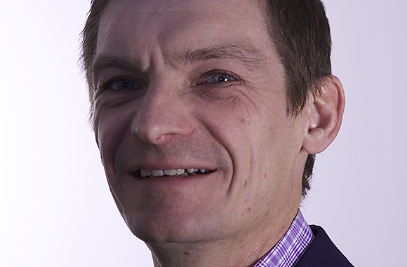 A close-up portrait of a middle-aged man with short brown hair smiling against a light background.