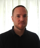 A man with short hair and a beard wearing a black polo shirt against a white curtain background.
