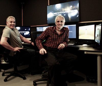 Two men smiling while sitting in front of multiple computer monitors in a dark production studio.