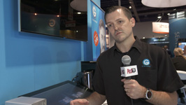 A man in a black polo shirt holds a microphone while standing in a convention hall.