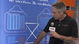 A man in a black polo shirt speaks into a microphone in front of a blue technical diagram.