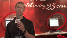 A man in a black polo shirt holds a microphone with a logo in front of a red patterned backdrop.