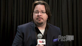 A man with glasses and a goatee wearing a dark blazer speaks into a microphone against a black curtain.