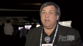 A man in a dark blazer and black shirt speaks into a silver microphone.