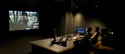 Two people sit at a desk with monitors in a dark room, watching a large projection screen.