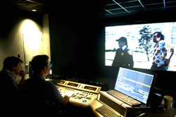 Two people sit at a desk with monitors and a control panel in a dark room, watching a large screen.