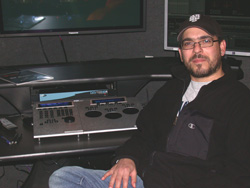 A man with glasses and a cap sits in front of monitors and a mixing console in a studio.