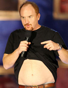 A man in a black t-shirt lifts his shirt to show his bare stomach while holding a microphone.