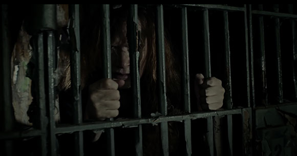 A person with long hair looks through dark metal prison bars in a dimly lit room.