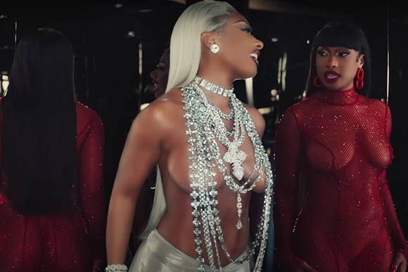 A woman in a silver jeweled harness looks at a woman in a red mesh bodysuit.