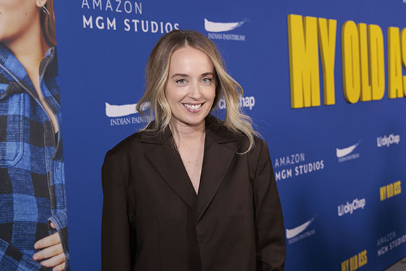 A woman with blonde hair wearing a dark blazer stands before a blue Amazon MGM Studios backdrop.