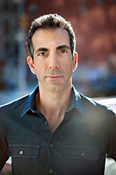 A man with dark hair and a dark button-down shirt looks directly at the camera against a blurred background.