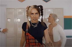 A woman with blonde and brown pigtails, wearing a plaid skirt and dark top, glares forward. Another person is visible behind