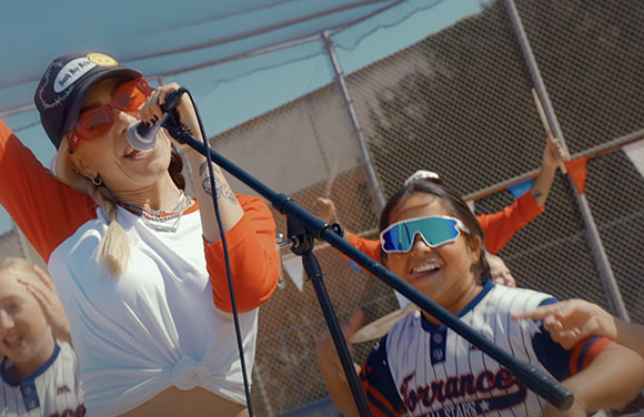 A woman sings into a microphone while a smiling person in a jersey stands behind her.