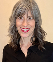 A woman with shoulder-length grey hair and a black shirt smiling against a neutral background.