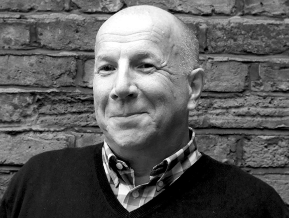 Black and white close-up portrait of a bald man smiling slightly against a brick wall background.