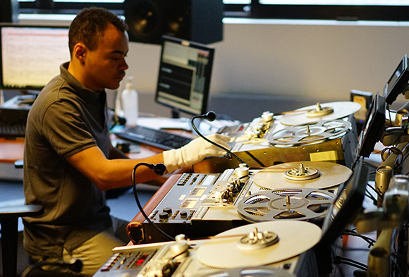 A man in a grey shirt uses white gloves to operate vintage reel-to-reel tape machines in a studio.